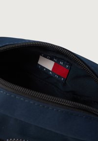 Interno di una borsa in tessuto blu navy con una zip nera; presenta un'etichetta con la scritta "TOMMY JEANS" accanto a un logo rosso, bianco e blu.