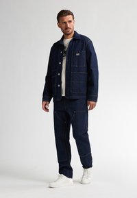 Uomo che indossa una giacca di jeans blu scuro e pantaloni, con sneaker bianche, in piedi e che guarda di lato su uno sfondo neutro.