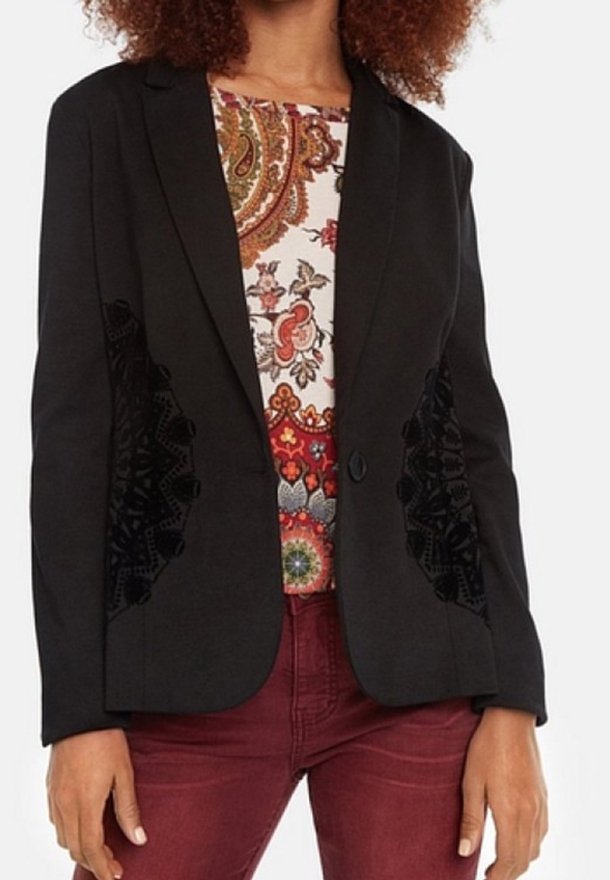 Mujer con un blazer negro con paneles laterales decorativos sobre una blusa colorida con estampado de cachemira y pantalones burdeos.
