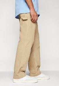 Beige wide-leg pants made of cotton, featuring a side pocket and visible stitching, paired with white sneakers.
