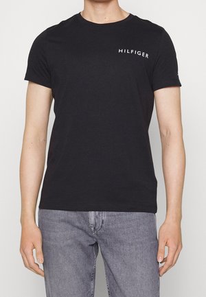 Man draagt een zwart T-shirt met ronde hals met de tekst "HILFIGER" op de borst, gecombineerd met lichtgrijze denim jeans, staand tegen een effen achtergrond.