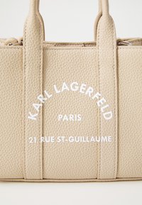 Beige texturerad handväska med två topphandtag, utrustad med präglad vit logotext på framsidan för Karl Lagerfeld, Paris.