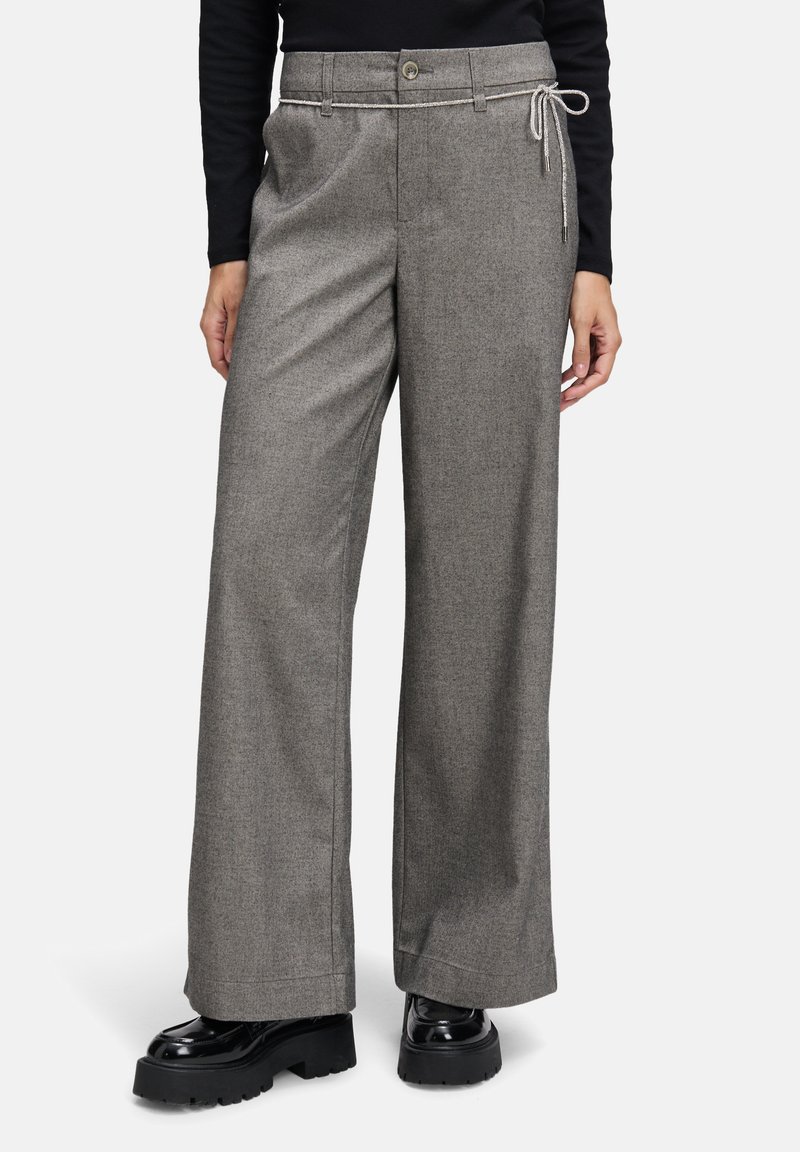Vrouw in een grijze wijde broek met een dunne touwriem en zwarte dikke platform schoenen, staand met de handen ontspannen langs de zij.