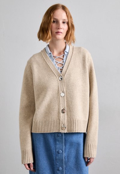 Cardigan bej din lână, cu guler în V, tiv ondulat și cinci butoane mari. Asortat cu o cămașă albastră, cu guler, care are un design cu șireturi.