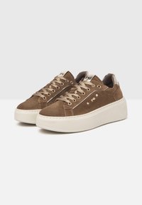 Sneaker in suede marrone con suola piatta bianca, occhielli dorati e borchie decorative dorate sui lati. Lacci piatti e supporto per la caviglia imbottito.