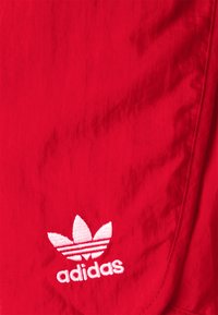 Červená sportovní tkanina s výrazným bílým logem Adidas trefoil, hladký povrch a čisté hrany.