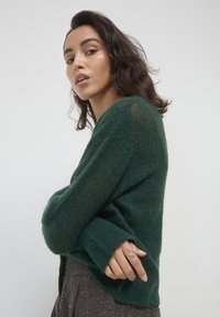 Dunkelgrüner, flauschiger Strickpullover mit gerippten Bündchen und lockerer Passform. Die Textur wirkt weich und wird durch einen stylischen Ring an der Hand des Models ergänzt.