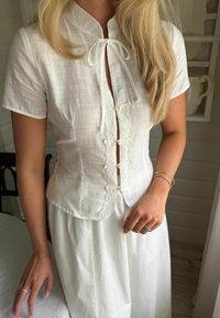 Femme portant un chemisier blanc à manches courtes avec dentelle et liens, associé à une jupe blanche, debout à l'intérieur près d'une chaise.