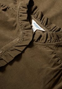 Olivos žalias viršus, pagamintas iš minkštos medžiagos, turintis raukšlėtą apykaklę, mygtukų užsegimus ir subtilią vertikalią raukšlėtą tekstūrą. Matomas prekės ženklas.