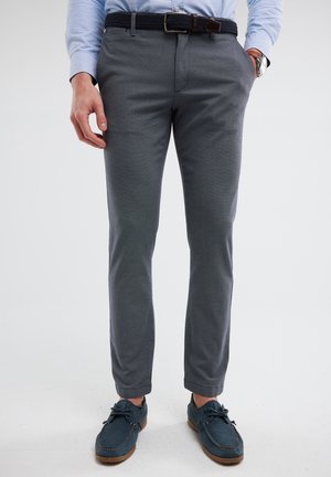 Pantalones gris claro de corte ajustado con una textura suave, que presentan bolsillos laterales y un cinturón negro trenzado. Combinados con zapatos azul con cordones.