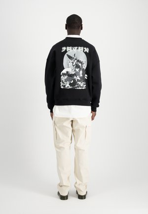Sort sweatshirt med hvid grafisk design på ryggen, der viser en karakter og bølger. Kombineret med lysebeige cargo bukser og en hvid skjorte.