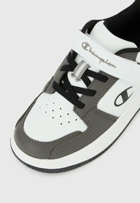 Λευκό και γκρί αθλητικό παπούτσι Champion με μαύρα κορδόνια, διάτρητη μύτη και ιμάντα βέλκρο με τη σήμανση "Champion" σε γραφή.