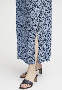 Blauwe maxi-jurk met zwart bladpatroon, heeft een zijsplit. Gepaard met zwarte instapsandalen, die een blokhakontwerp laten zien.