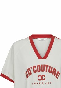 Balta marškinėliai su raudonu V formos iškirpimu ir raudonais akcentais, su užrašu "GO'COUTURE LOVE & JOY" ryškiomis raudonomis raidėmis per visą priekį.
