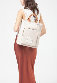 Donna che indossa una gonna color ruggine e una maglietta bianca, con una borsa tote beige con manici marroni e una tasca con zip frontale sulla schiena.