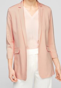 Vaaleanpunainen bleiseri, jossa on istuva leikkaus, lovettu kaulus ja kaksi etutaskua. Päällä vaaleanpunainen V-pääntiestoppi ja valkoiset housut.