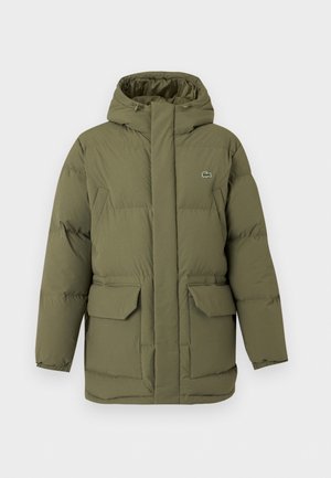 Homme portant une veste matelassée vert olive, un t-shirt blanc, un jean bleu et des baskets blanches, debout devant un fond blanc uni.