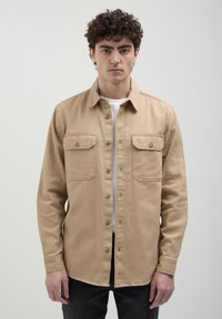 Beige Hemd mit Knopfleiste aus robustem Material, ausgestattet mit zwei Brusttaschen und einem kragenlosen Design, kombiniert mit einem weißen T-Shirt darunter.
