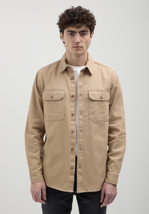 Beige Hemd mit Knopfleiste aus robustem Material, ausgestattet mit zwei Brusttaschen und einem kragenlosen Design, kombiniert mit einem weißen T-Shirt darunter.