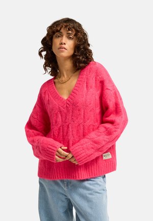 Donna con capelli ricci che indossa un maglione rosa acceso lavorato a trecce e jeans azzurri, in piedi davanti a uno sfondo semplice.