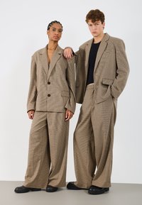 Blazere oversized în carouri maro, cu pantaloni largi dintr-un material texturat, având două butoane și buzunare frontale. Asortate cu încălțăminte neagră.
