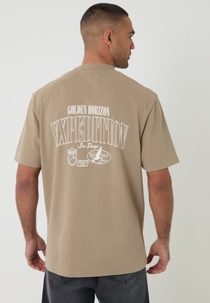 Uomo con maglietta beige con la scritta "Golden Horizon Expedition San Diego" e grafica a tema marittimo sul retro, in piedi davanti a uno sfondo neutro.