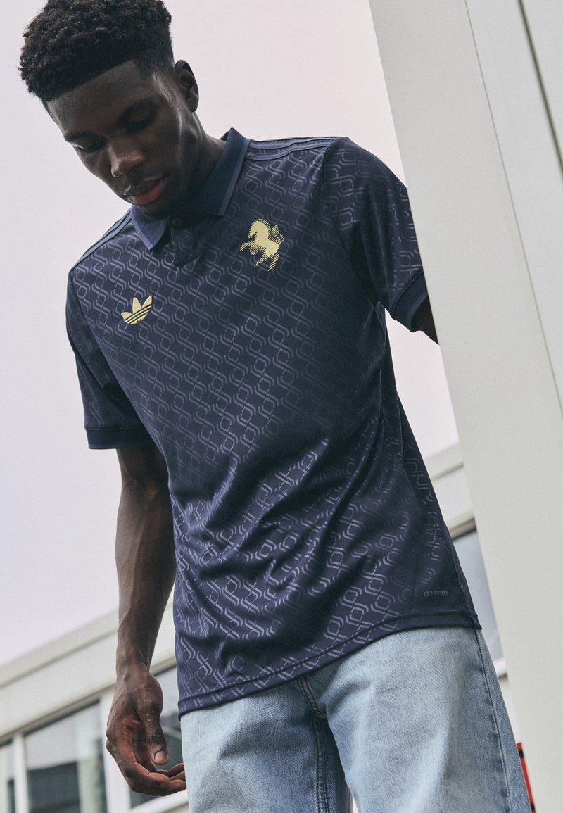 Marineblaues Poloshirt mit geometrischem Muster, Polo-Kragen und goldenem Löwenlogo. Kombiniert mit hellblauen Jeans, wird das Modell von oben aufgenommen.