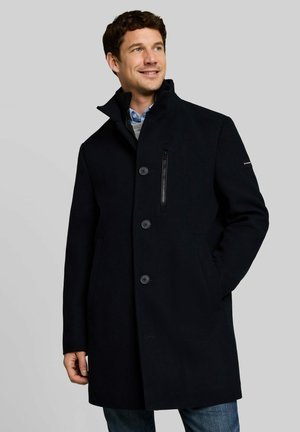 Uomo che indossa un cappotto nero con bottoni e collo alto, camicia blu e maglione grigio sotto, mani nelle tasche, guarda leggermente a destra e sorride.