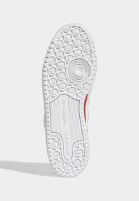 Semelle de chaussure Adidas blanche avec des motifs de bande de roulement circulaires et en forme d’étoile, avec le logo Adidas au centre, vue de dessous sur un fond clair.