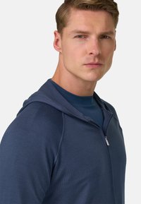 Sweat à capuche zippé bleu marine avec un tissu texturé, doté d'une capuche et d'une chemise bleue contrastante en dessous. Vue en gros plan du profil du modèle.