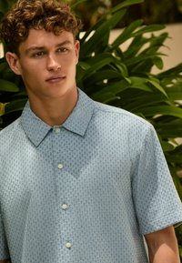Joven con cabello rizado que lleva una camisa de manga corta color azul claro con estampado y botones, de pie frente a plantas verdes con hojas.