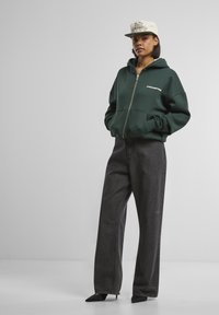 Grøn zip-hoodie med frontlommer, parret med højtaljede mørke denimjeans og en beige caps med dekorativ tekst.