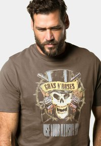 Brun bomulds T-shirt med et grafisk design af et kranium i en hat, flankeret af krydsede våben og teksten "GUNS N' ROSES USE YOUR ILLUSION."