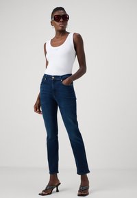 Un débardeur blanc à côtes, un jean droit bleu foncé avec des accents en bouton doré, et des mules noires à talons. De grandes lunettes de soleil carrées complètent le look.