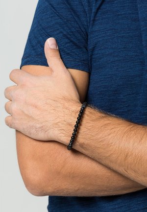 Schwarzes Perlenarmband an einem Handgelenk, mit runden Perlen in glatter Textur. Über einem blauen Stoffhemd getragen, was einen lässigen Look zeigt.
