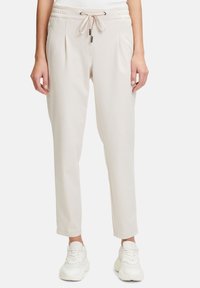 Lichtbeige joggerpants met een trekkoord in de taille, geplooide voorkant, taps toelopende benen en een gladde textuur, gecombineerd met witte sneakers.