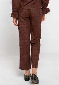 Persona con pantalones a cuadros marrones y blusa a juego con mangas con volantes, combinados con zapatos de tacón bajo negros, de pie sobre un suelo blanco.