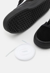 Sneaker nera con suola in gomma spessa e cuciture bianche accanto a un laccio di scarpa bianco arrotolato su una superficie bianca.