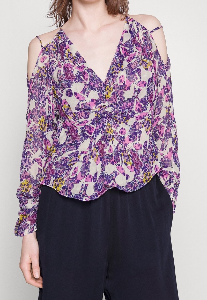 Femme portant un chemisier à épaules dénudées à motifs floraux violet, rose, jaune et blanc avec un devant froncé, associé à un pantalon noir ample.