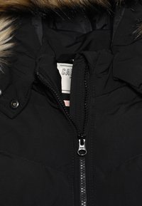 Černá bunda s kapucí na zip, která má kožíškový lem na kapuci a strukturovanou látku. Pozoruhodný detail se stříbrným zipem.