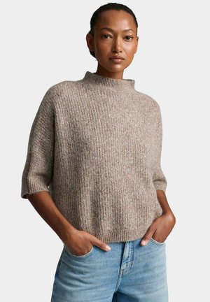 Femme aux cheveux courts portant un pull en maille beige ample avec des manches aux coudes et un jean en denim bleu clair, les mains dans les poches.