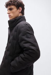 Μαύρο γιλέκο με ψηλό γιακά, διαθέτει λεία υφή, πλαϊνές τσέπες και ετικέτα στον άνω βραχίονα. Εμφανίζεται πλαϊνή όψη.