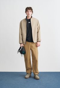 Μπεζ ζακέτα με φερμουάρ πάνω από μαύρο T-shirt, μαύρο παντελόνι, γκρι μπότες· κρατώντας μια μικρή μαύρη τσάντα, στέκεται σε μπλε πάτωμα μπροστά σε λευκό τοίχο.