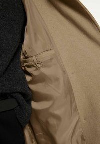 Beige ytterkläder med en slät satinliknande foder, innehållande en liten ficka och mörk texturerad tyg synlig på vänster sida.