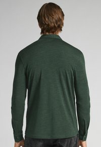 Mörkgrön långärmad skjorta, tillverkad av en texturerad tyg. Har en krage och knäppta ärmslut, med en slät, figurnära bakdesign.