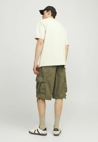 Beżowa koszula z krótkim rękawem w połączeniu z oliwkowymi szortami typu cargo z bocznymi kieszeniami. Na nogach białe sneakersy i beżowe skarpetki. Na głowie czarna czapka.