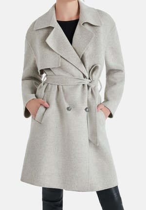 Femme portant un manteau en laine gris clair ceinturé avec de larges revers et des détails de boutons, mains dans les poches, sur un pantalon et un haut noirs.