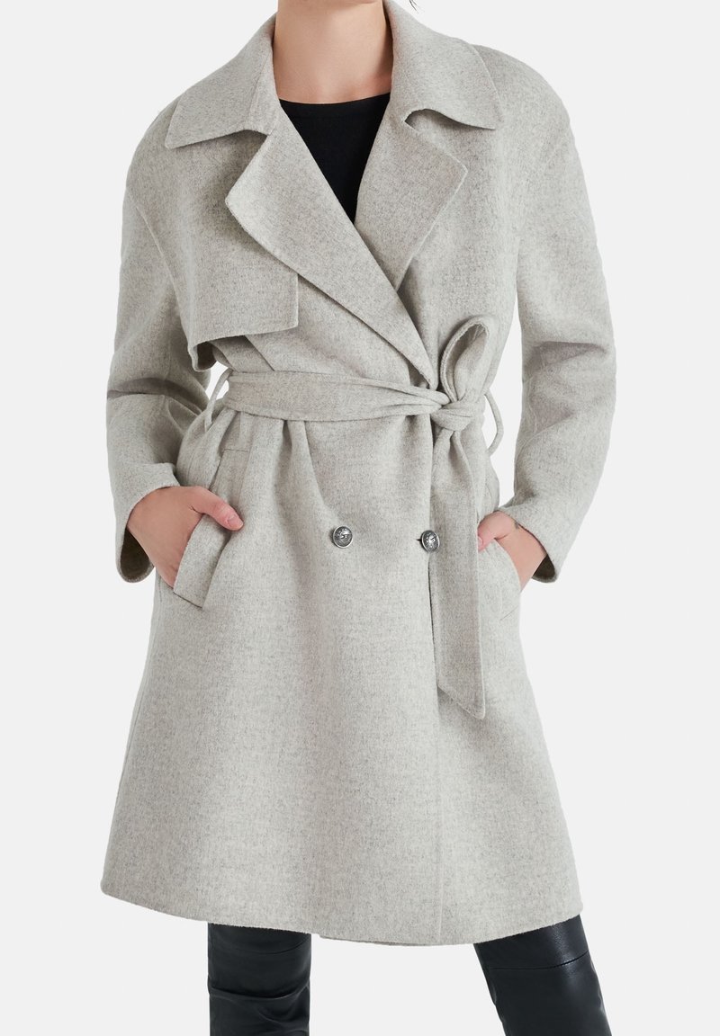 Femme portant un manteau en laine gris clair ceinturé avec de larges revers et des détails de boutons, mains dans les poches, sur un pantalon et un haut noirs.
