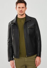 Giacca in pelle nera con zip frontale, tasche laterali e tasca sul petto. Presenta un colletto e una vestibilità sagomata. Indossata sopra un pullover verde.