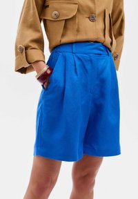 Blauwe katoenen shorts met plooien, een ontspannen pasvorm en zijzakken. Gestyled met een bruine shirt en een bordeauxrode armband om de pols.